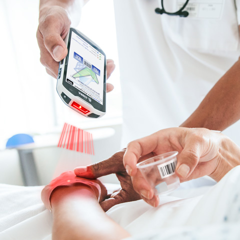 Mobiles Datenerfassungsgerät im Smart Design, mit dem am Krankenhausbett ein Patientenarmband gescannt wird.